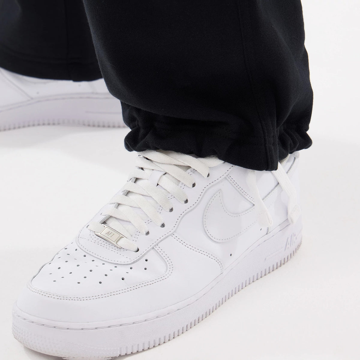 New Era Black Straight Legged Joggers, close-up view showing the elasticated cuffs resting over white sneakers, highlighting the smooth black fabric and loose fit around the ankles.