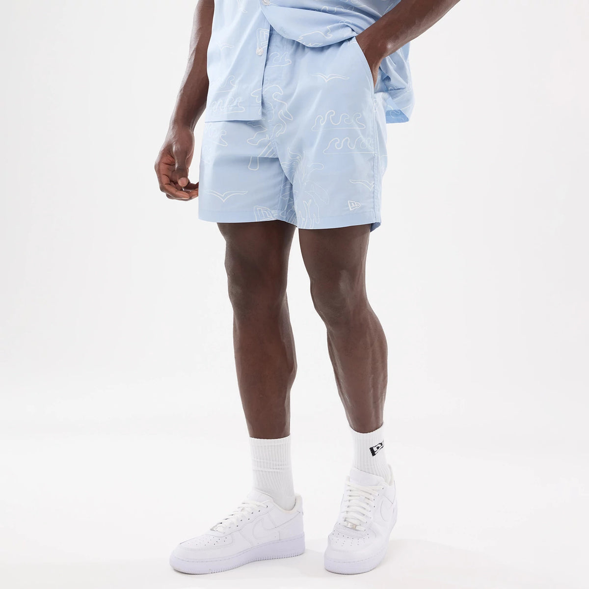 New Era Resort Blue Oversized Shorts, on-model view showing the light blue fabric with white abstract patterns, a raised New Era logo on the lower left leg and an elastic waistband for a relaxed fit.