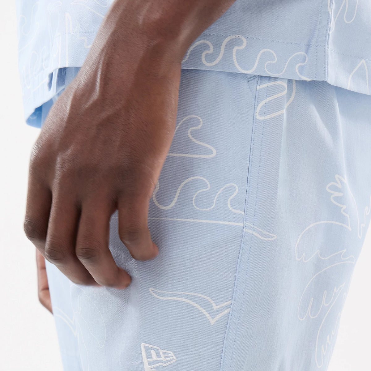 New Era Resort Blue Oversized Shorts, side view showing the light blue fabric with wave and abstract patterns, a debossed New Era logo near the hem, and a hand resting lightly on the side of the shorts.