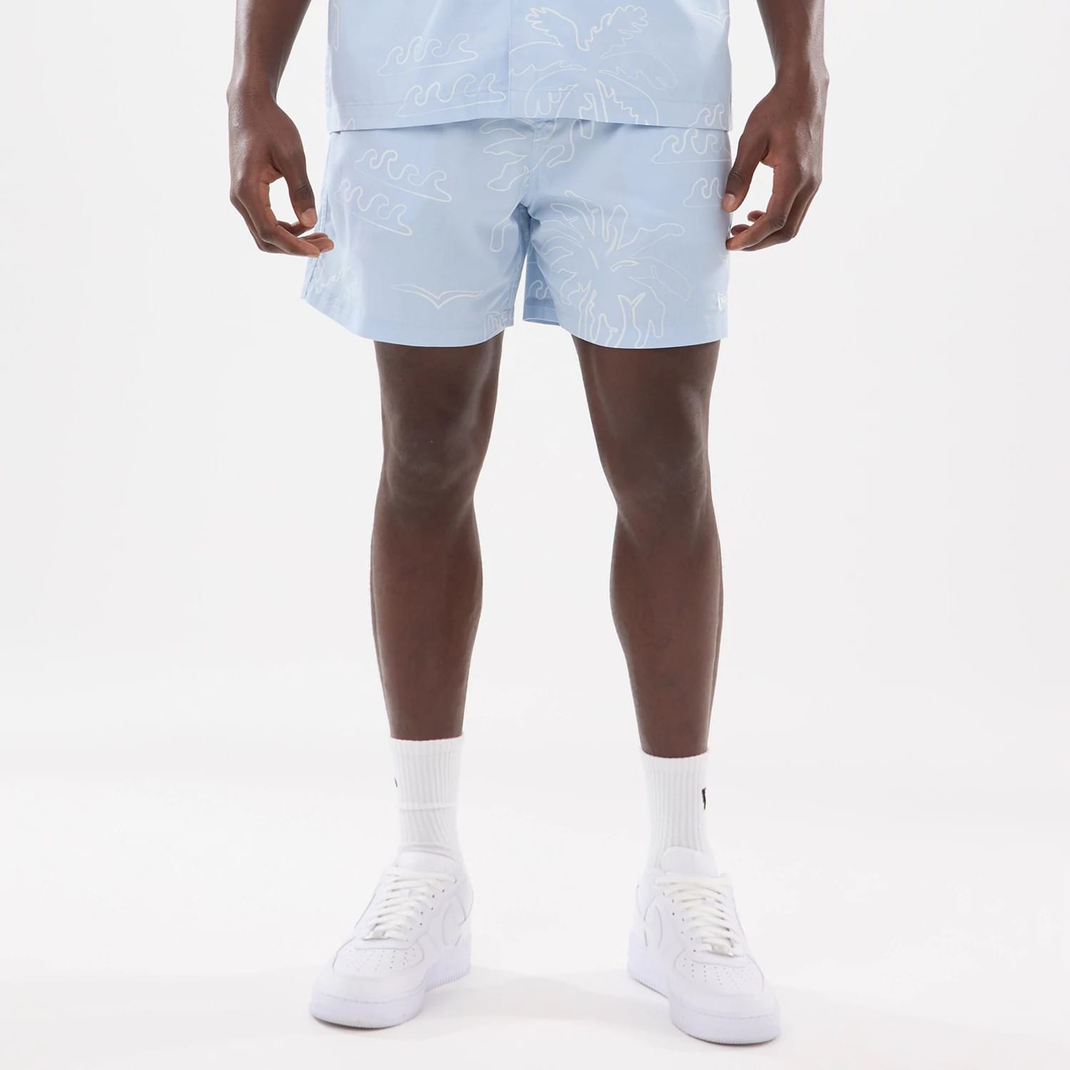 New Era Resort Blue Oversized Shorts, front view showing light blue shorts with white wave and palm tree patterns, reaching mid-thigh, worn with a matching blue shirt, and white sneakers completing the outfit.