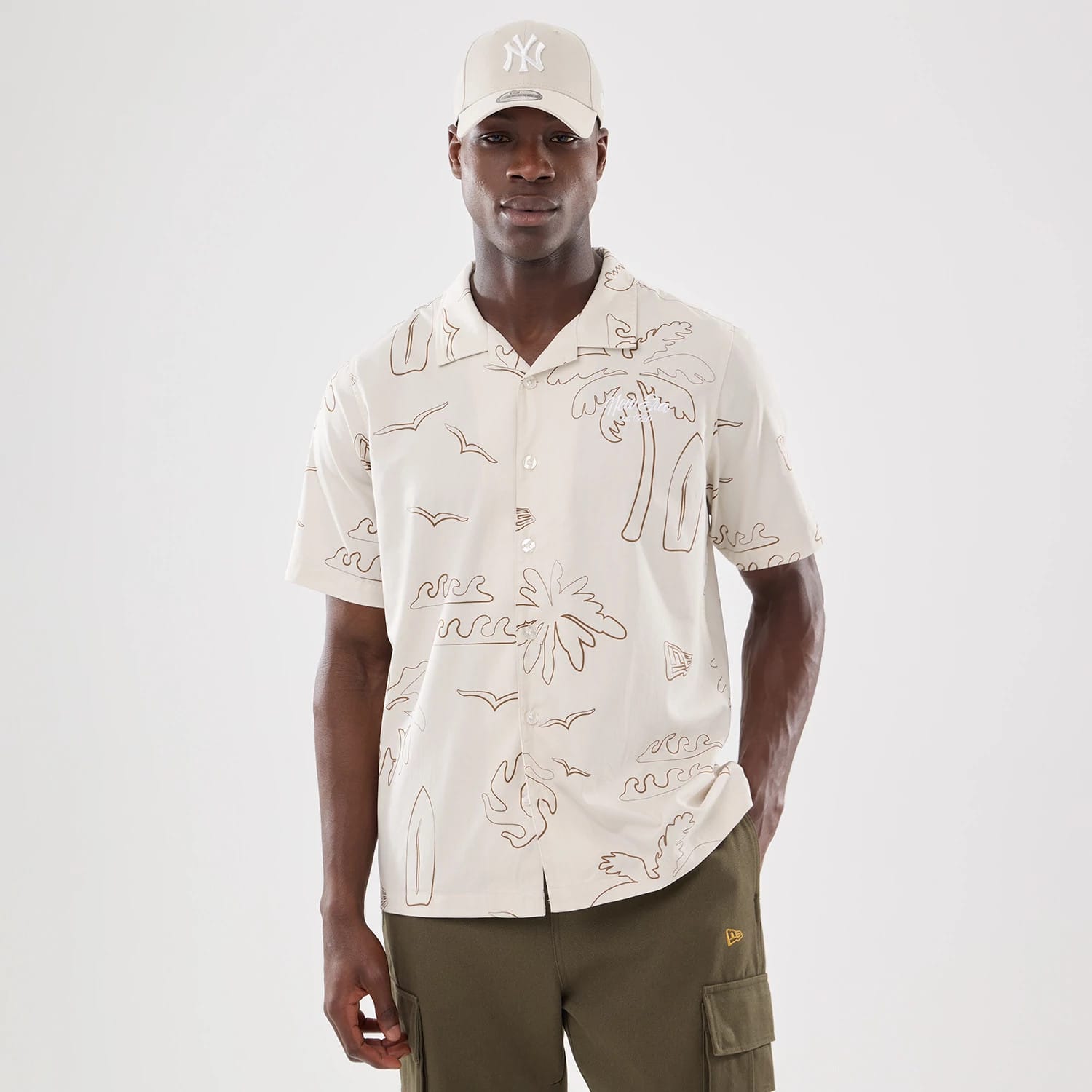 New Era Resort Natural Short Sleeve Shirt, front view on a model paired with a natural New York Yankees 9FORTY cap, featuring brown line illustrations of palm trees, birds, and surfboards on the shirt.