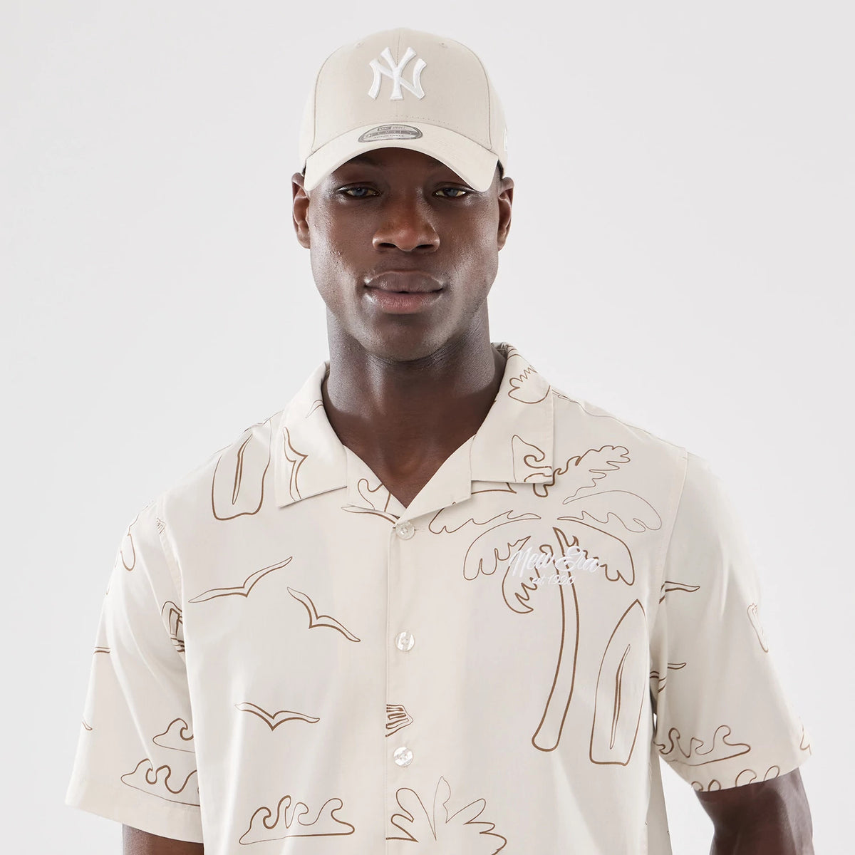 New Era Resort Natural Short Sleeve Shirt, front view on a model wearing a matching cap, featuring a natural colour with tropical line art motif, including palm trees and waves, on a relaxed fit with a button collar.