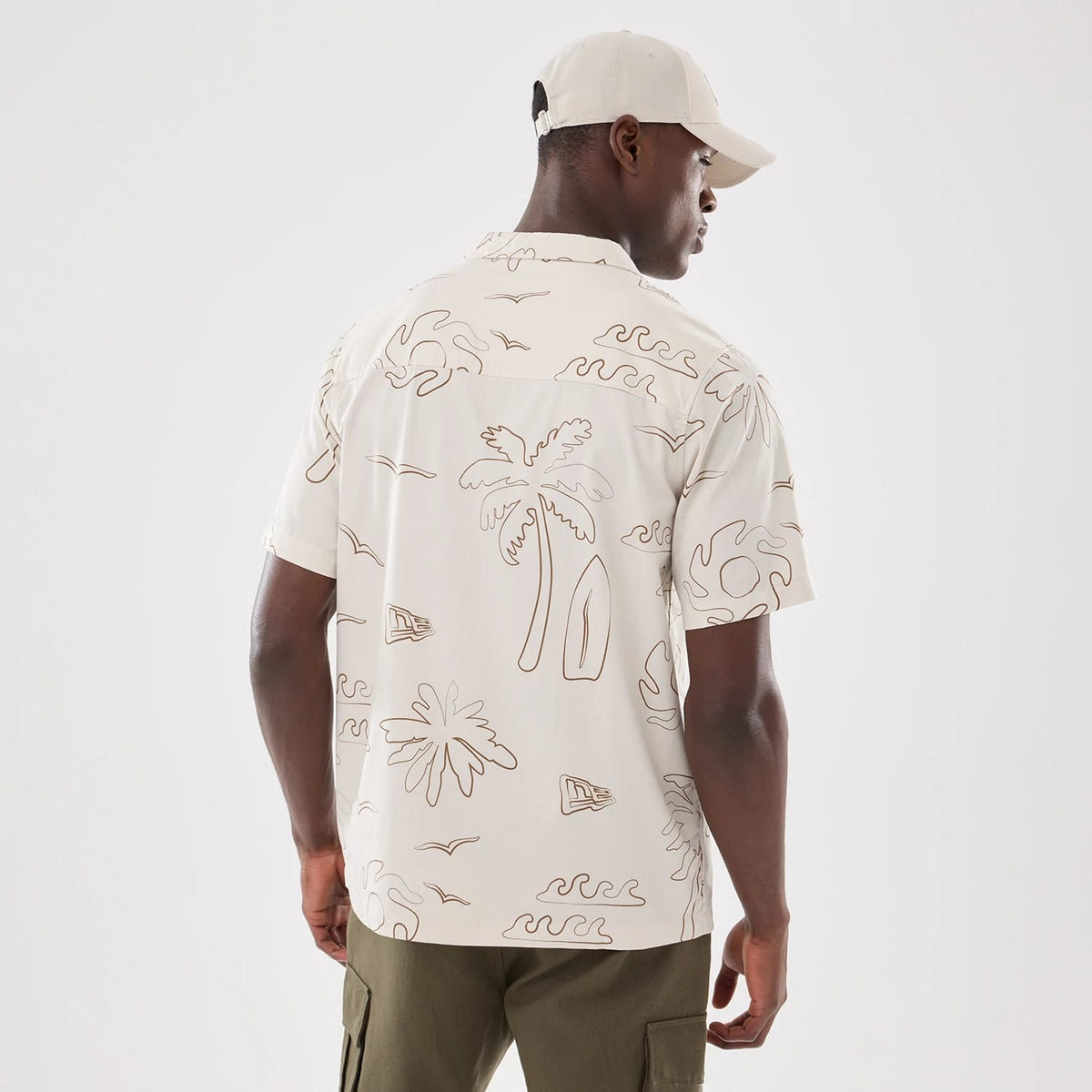 New Era Resort Natural Short Sleeve Shirt, back view on a model wearing a light cap, features a brown line-drawn pattern of palm trees, surfboards, and waves, covering the ivory fabric and extending across the back.