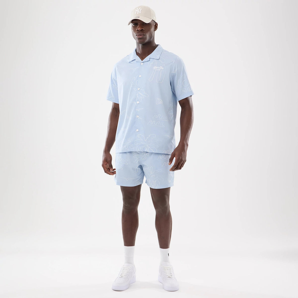 New Era Resort Blue Short Sleeve Shirt, front view on a model wearing matching shorts and a beige cap, featuring a light blue colour with white abstract line patterns and white buttons down the front.