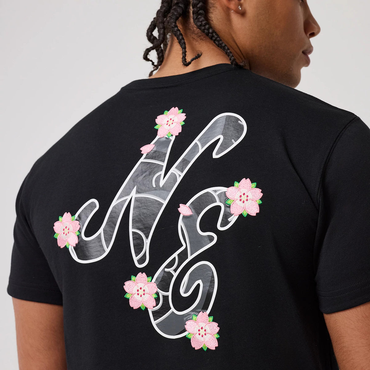 New Era Sakura Black T-Shirt, rear view showing a large white NE logo overlaid with cherry blossom designs across the back, worn by a person, the pink flowers adding a contrasting detail to the black fabric.