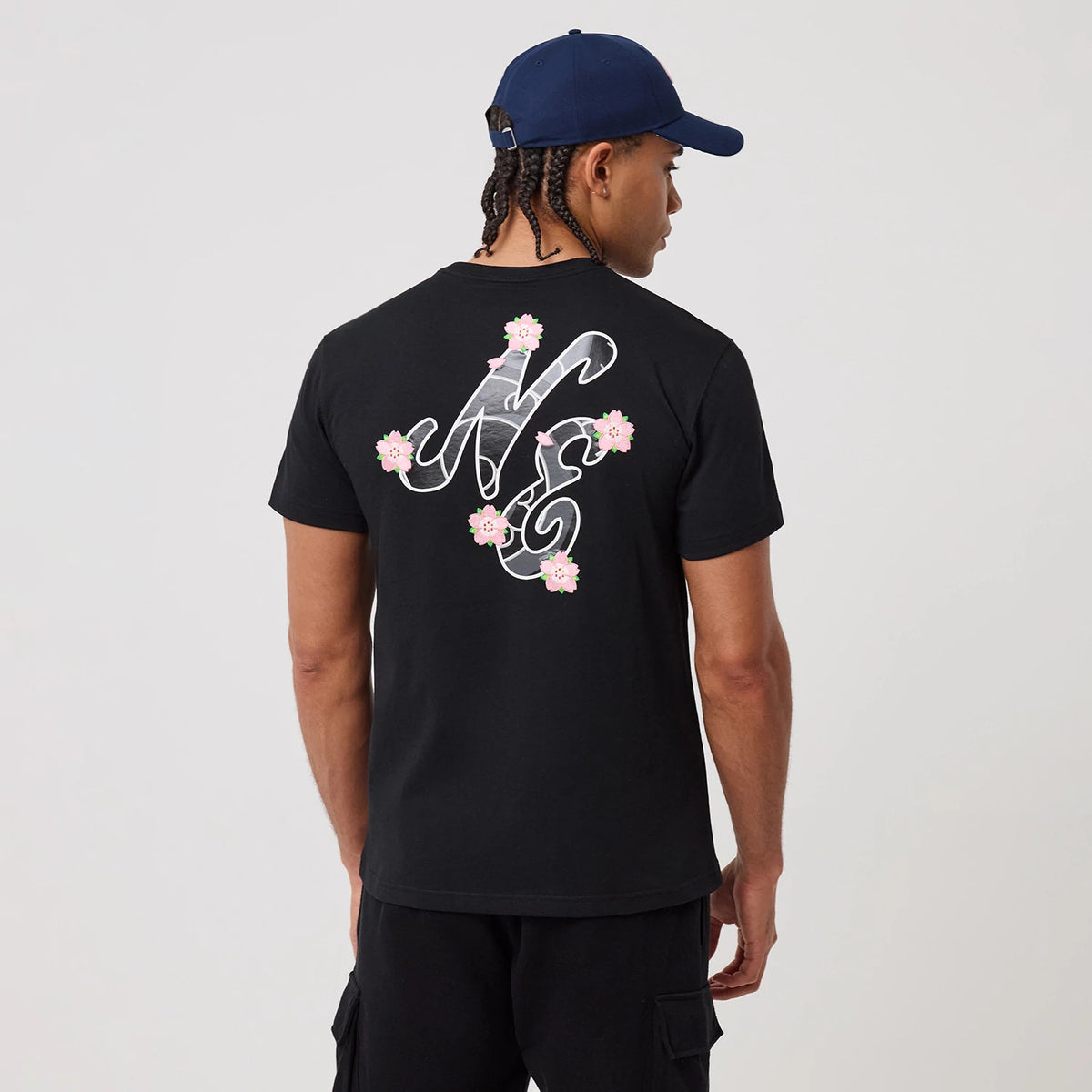 New Era Sakura Black T-Shirt, rear view displaying a large stylized NE graphic on the upper back adorned with pink cherry blossom designs, worn by a male model in black shorts and a navy cap.