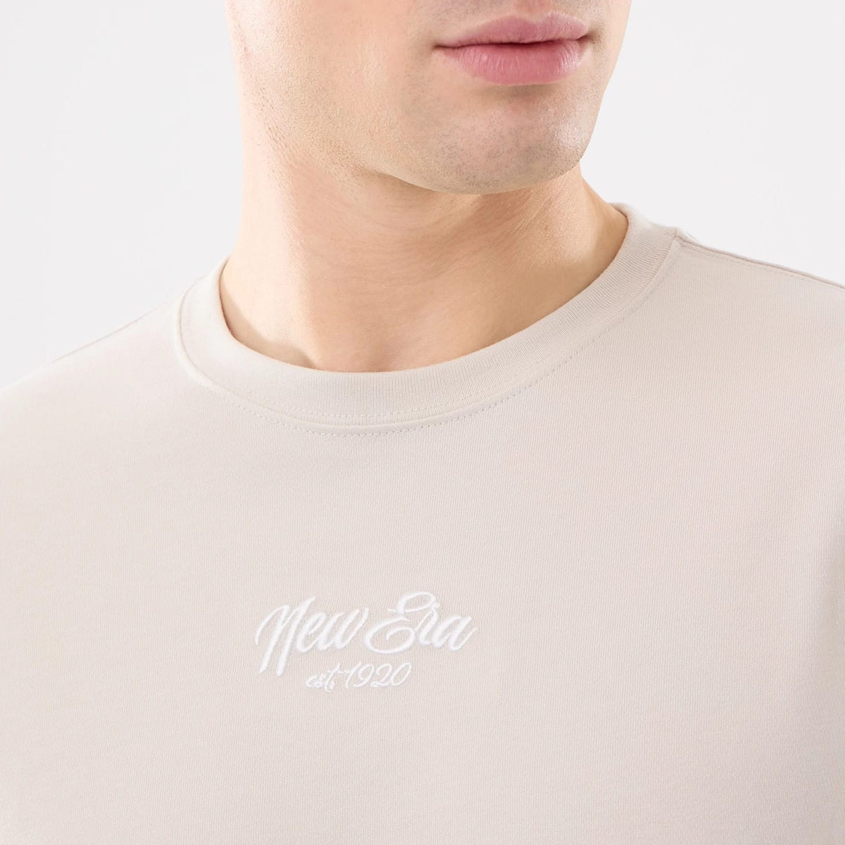 New Era Resort Natural T-Shirt, on-model view showing the upper section of a light cream t-shirt with a raised white New Era logo and "est. 1920" text centred on the chest below the round neckline.