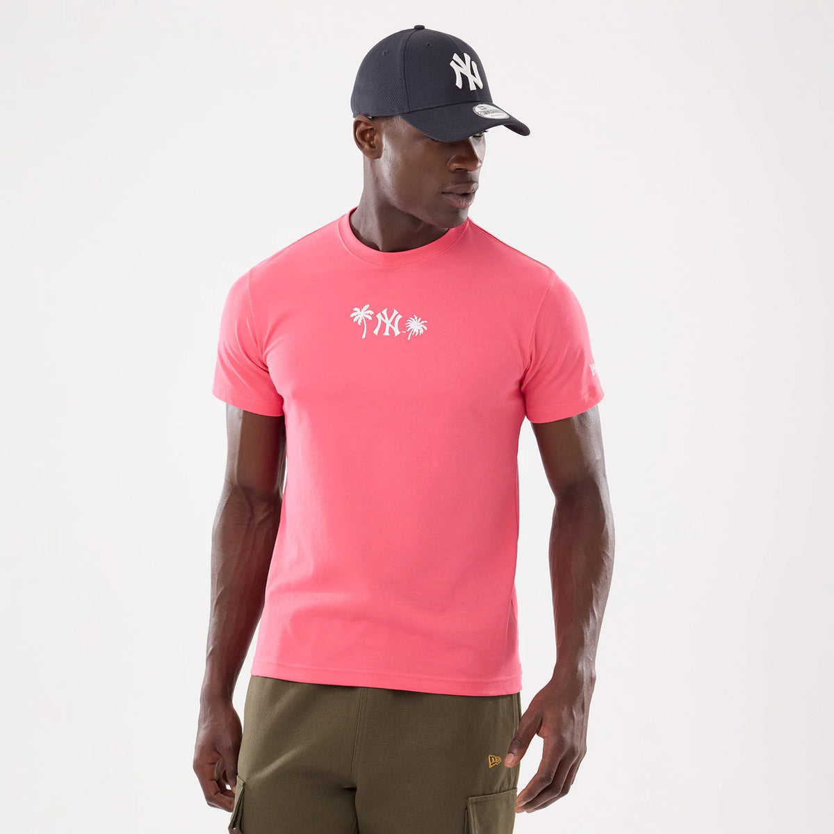 New York Yankees MLB Resort Pink T-Shirt, front view showing the bright pink fabric with a printed white Yankees logo between two palm trees across the chest, worn by a model in olive green trousers and a dark cap.