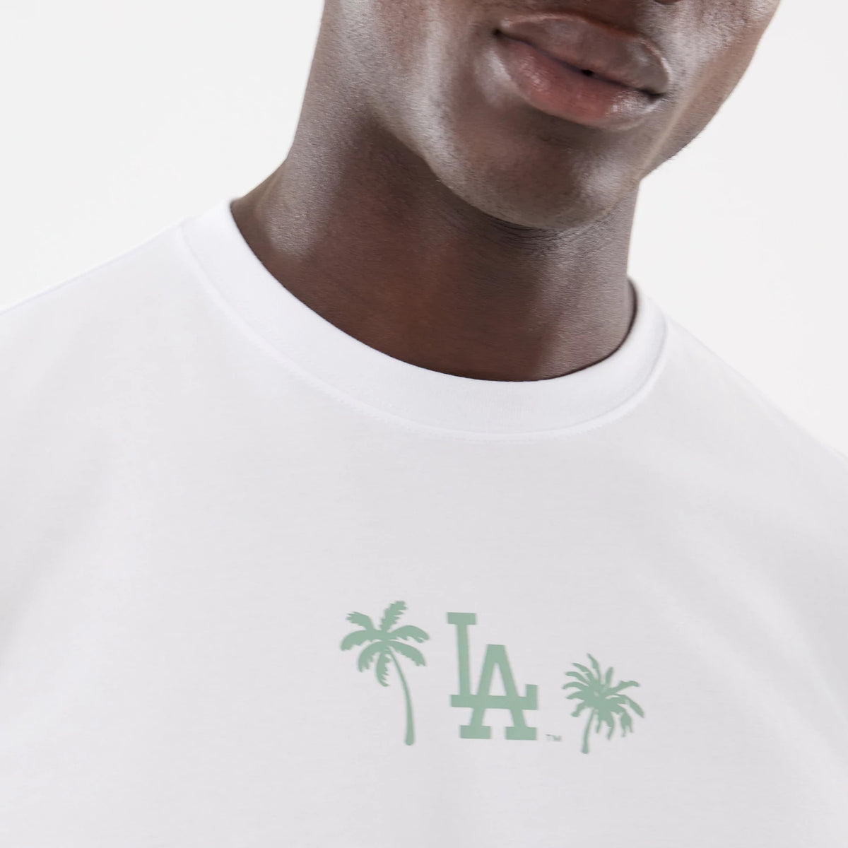 New Era LA Dodgers MLB Resort White T-Shirt, on-model view showing a close-up of the chest area with a green debossed LA logo flanked by palm tree graphics on a white fabric backdrop.