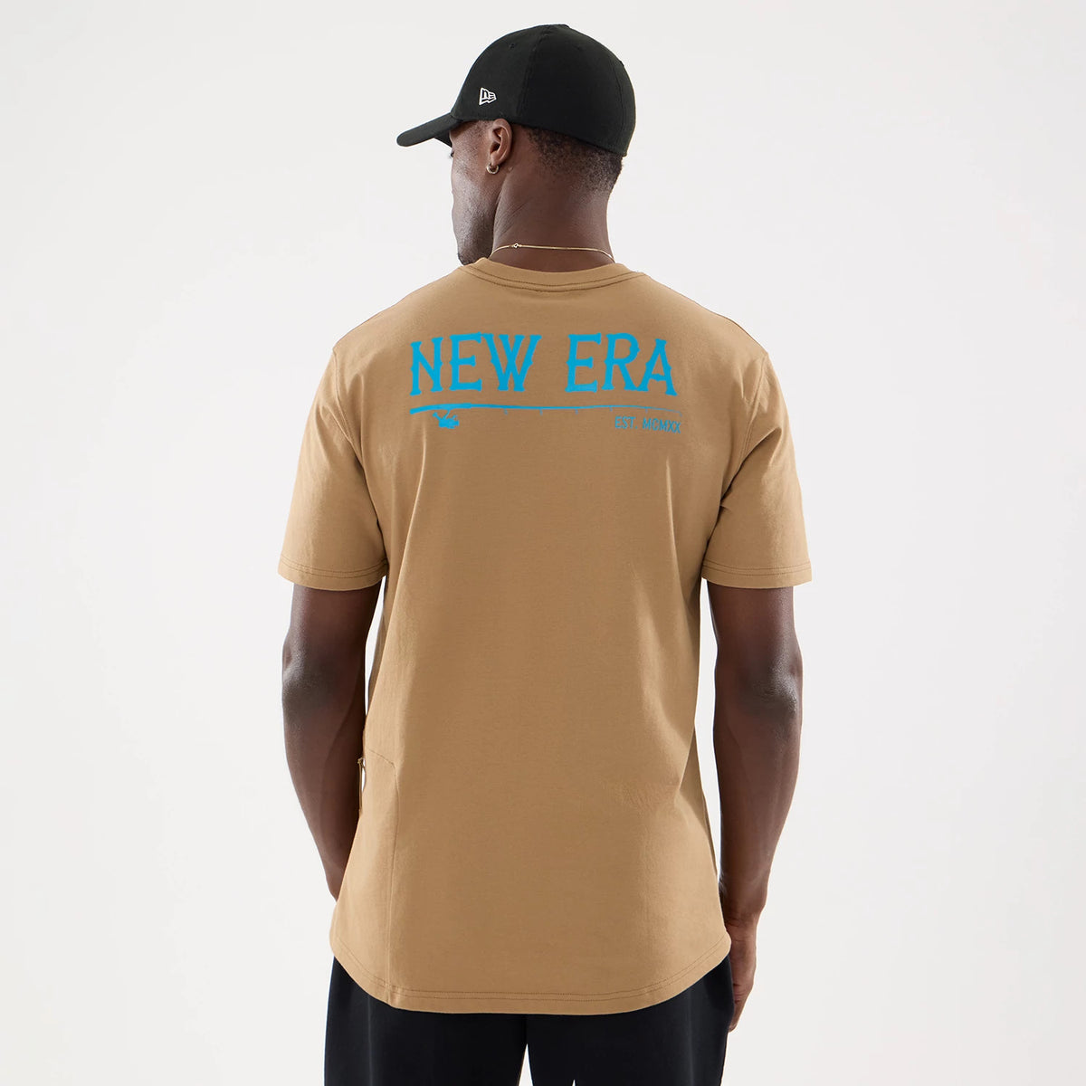 New Era Graphic Outdoor Fishing Beige T-Shirt, rear view showing the blue graphic text "NEW ERA" in large lettering across the upper back, worn by a model facing away, paired with a black cap featuring a raised New Era logo on the side.