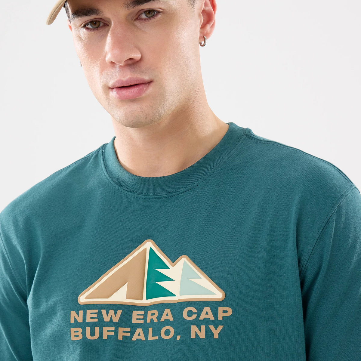 New Era Outdoor Mountain Dark Green T-Shirt, front view showing the graphic design with stylized mountains in beige and teal above the text "NEW ERA CAP BUFFALO, NY" printed in beige across the chest.