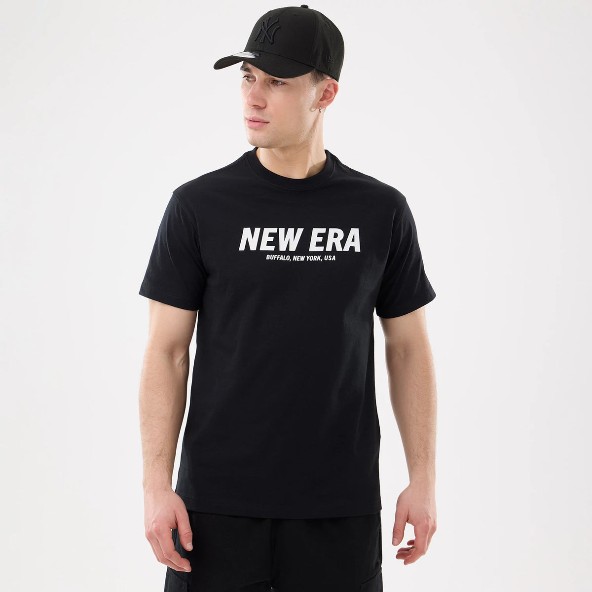 New Era Basic Black T-Shirt, on-model view showing a black t-shirt with a large white New Era logo centred on the chest, worn by a person also wearing a black cap with a raised logo.
