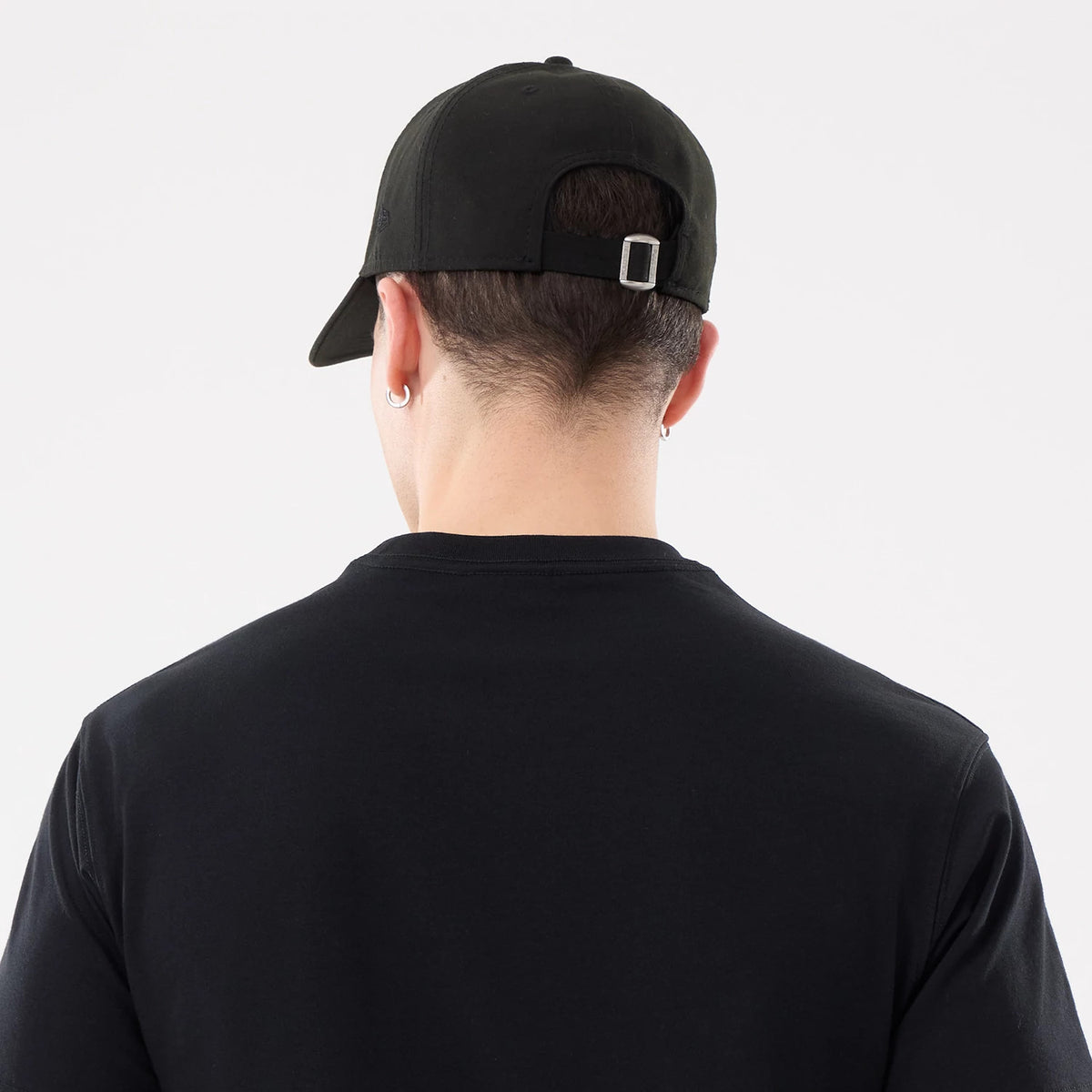 New Era Basic Black T-Shirt, rear view showing the plain black fabric and the back of a black cap worn, featuring a silver buckle adjustment and eyelets on the cap, worn by a person standing with their back to the camera.