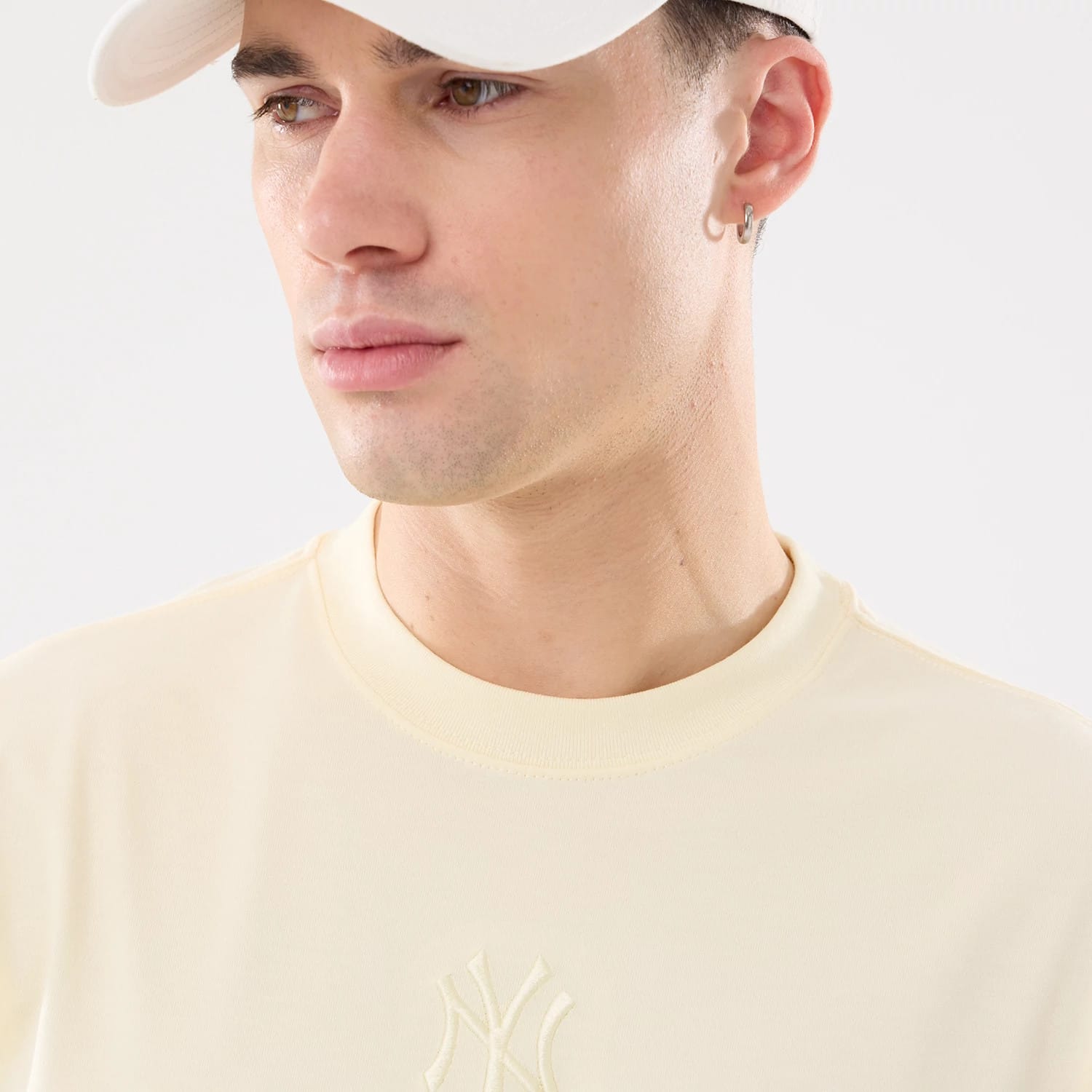 New Era New York Yankees MLB Cartoon Cream T-Shirt, on-model view showing the cream fabric with a raised NY Yankees logo centred on the chest, a round neckline and a white cap worn by the model.