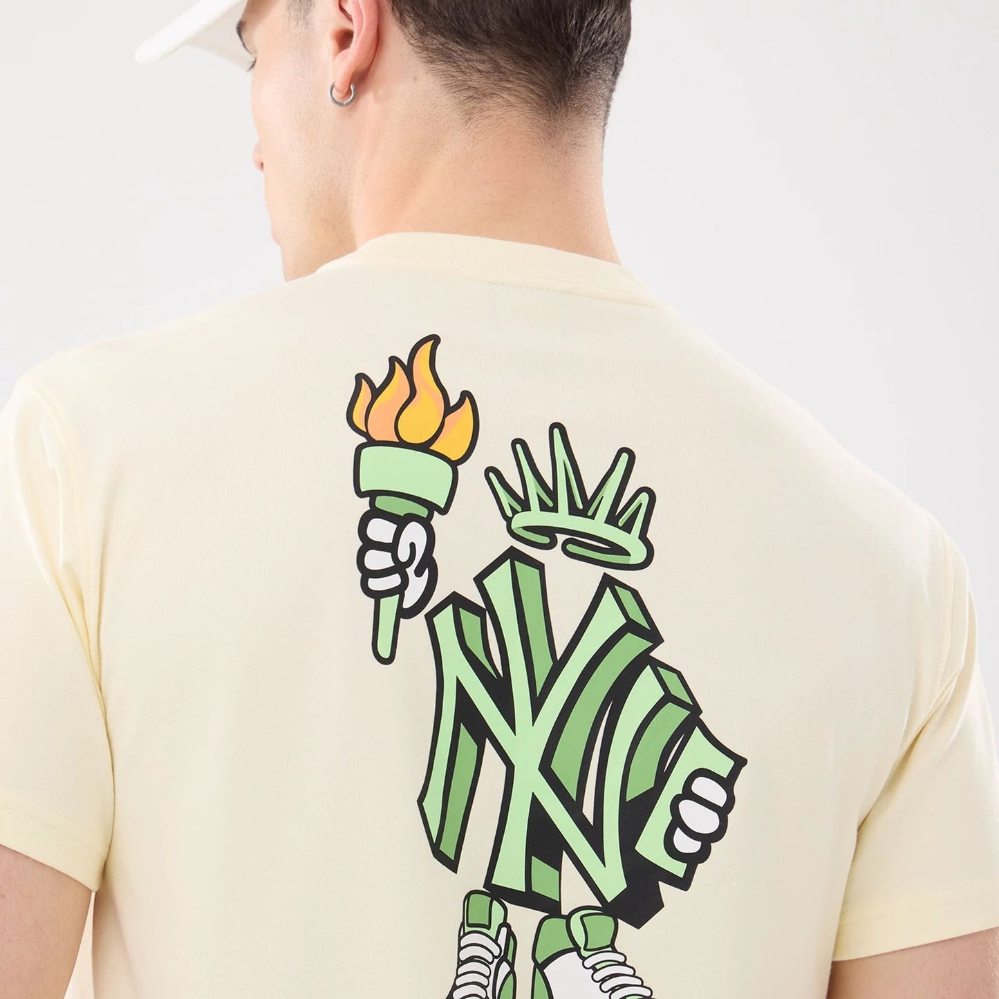 New York Yankees MLB Cartoon Cream T-Shirt, rear view showing a cartoon graphic of a green torch-bearing figure with a crown and sneakers, incorporating the Yankees logo on a cream background.