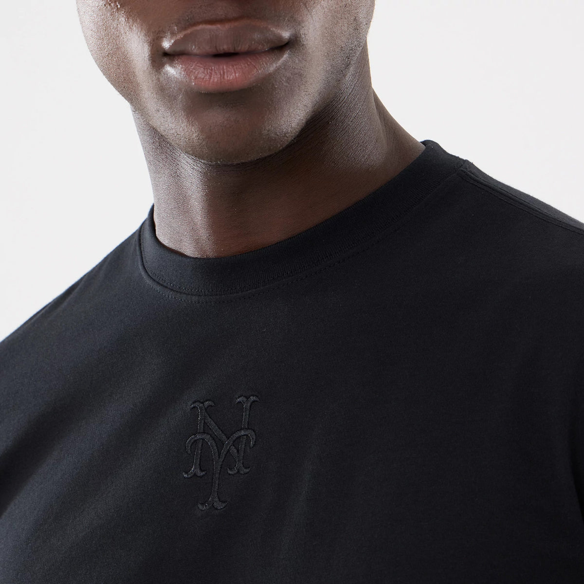New York Mets MLB Cartoon Black T-Shirt, front view showing the black cotton fabric with an embroidered New York Mets logo in black thread positioned on the left side of the chest.