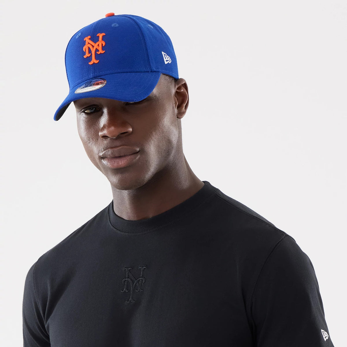 New York Mets MLB Cartoon Black T-Shirt, on-model view showing the black top with a raised black Mets logo on the front, paired with a blue cap featuring an orange Mets logo on the front and a white raised New Era logo on the side.
