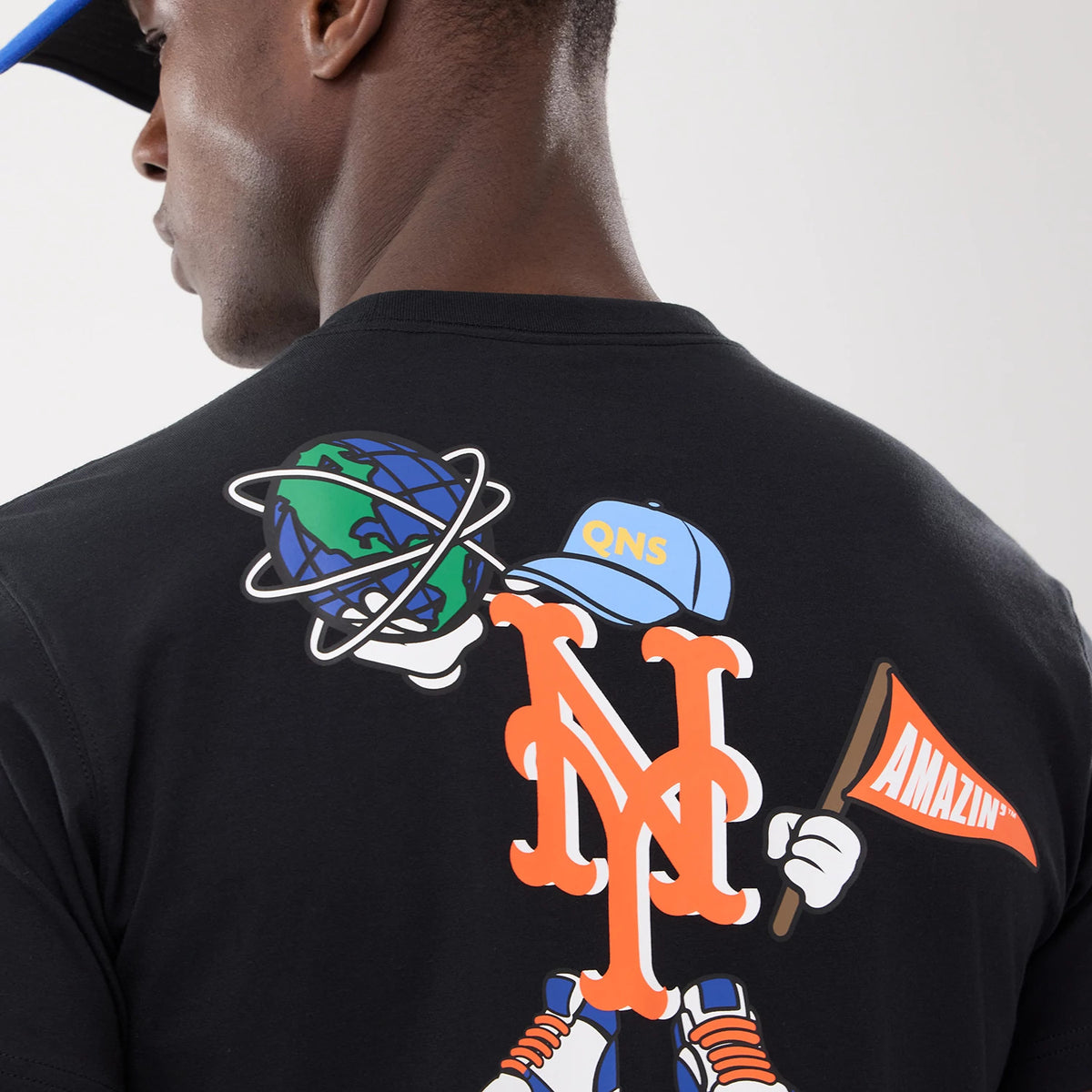 New Era New York Mets MLB Cartoon Black T-Shirt, rear view showing a large orange and white NY Mets logo with a cartoon character holding a globe and a flag, wearing a cap with QNS on it, printed on the black fabric.