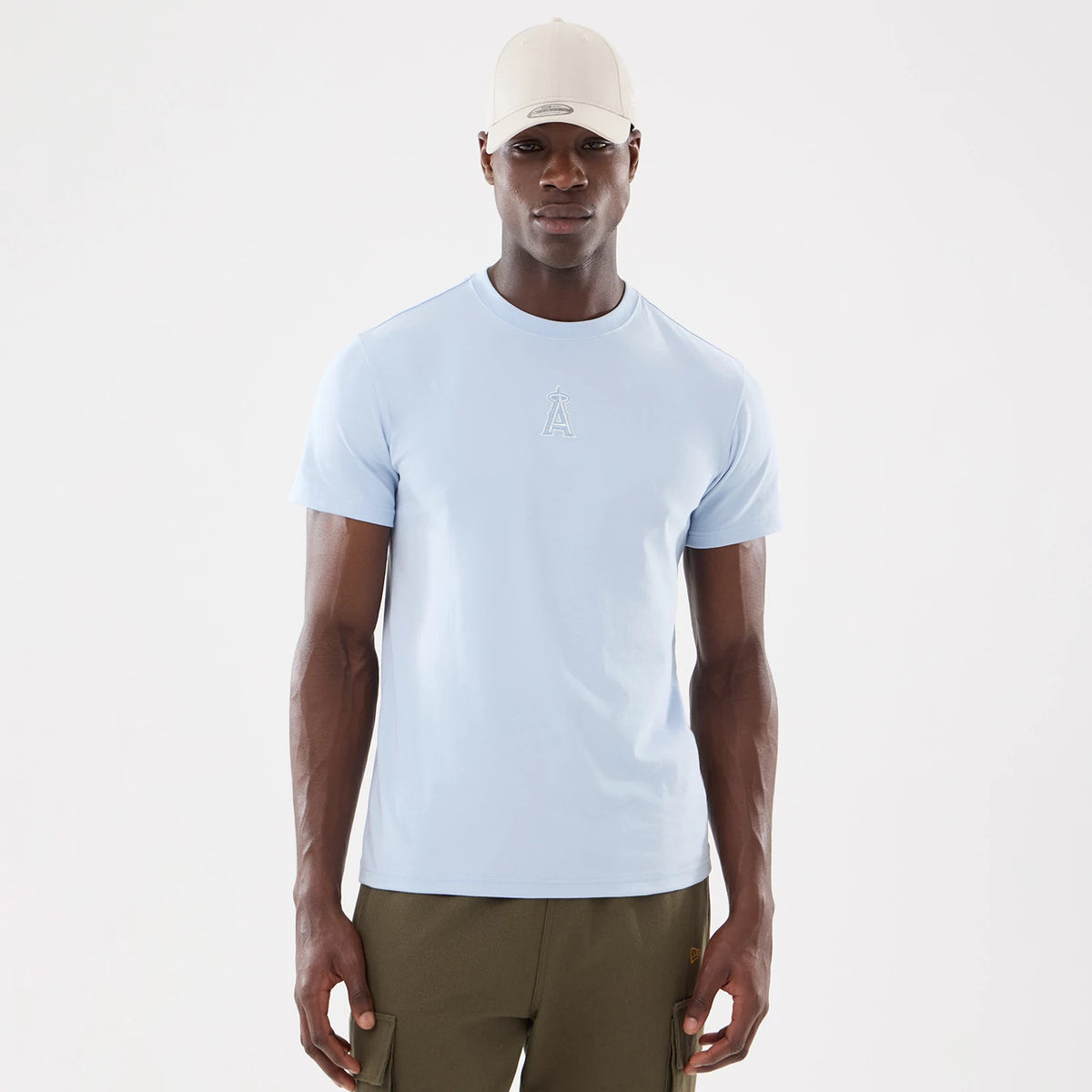 New Era LA Angels MLB Cartoon Blue T-Shirt, front view showing a light blue T-shirt with a raised LA Angels logo centred below the neckline, worn by a model also wearing a beige cap and olive green trousers.