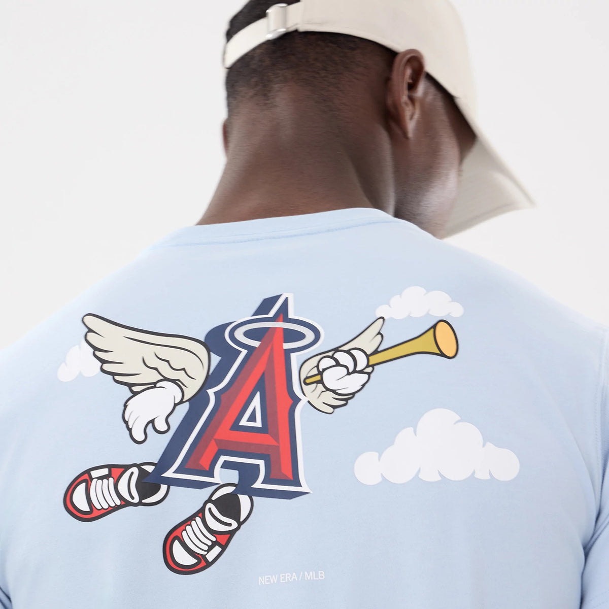 LA Angels MLB Cartoon Blue T-Shirt, on-model view showing the back of a light blue t-shirt with a large red and blue cartoon logo featuring wings and trumpet, with small clouds, worn by a person with a beige cap.