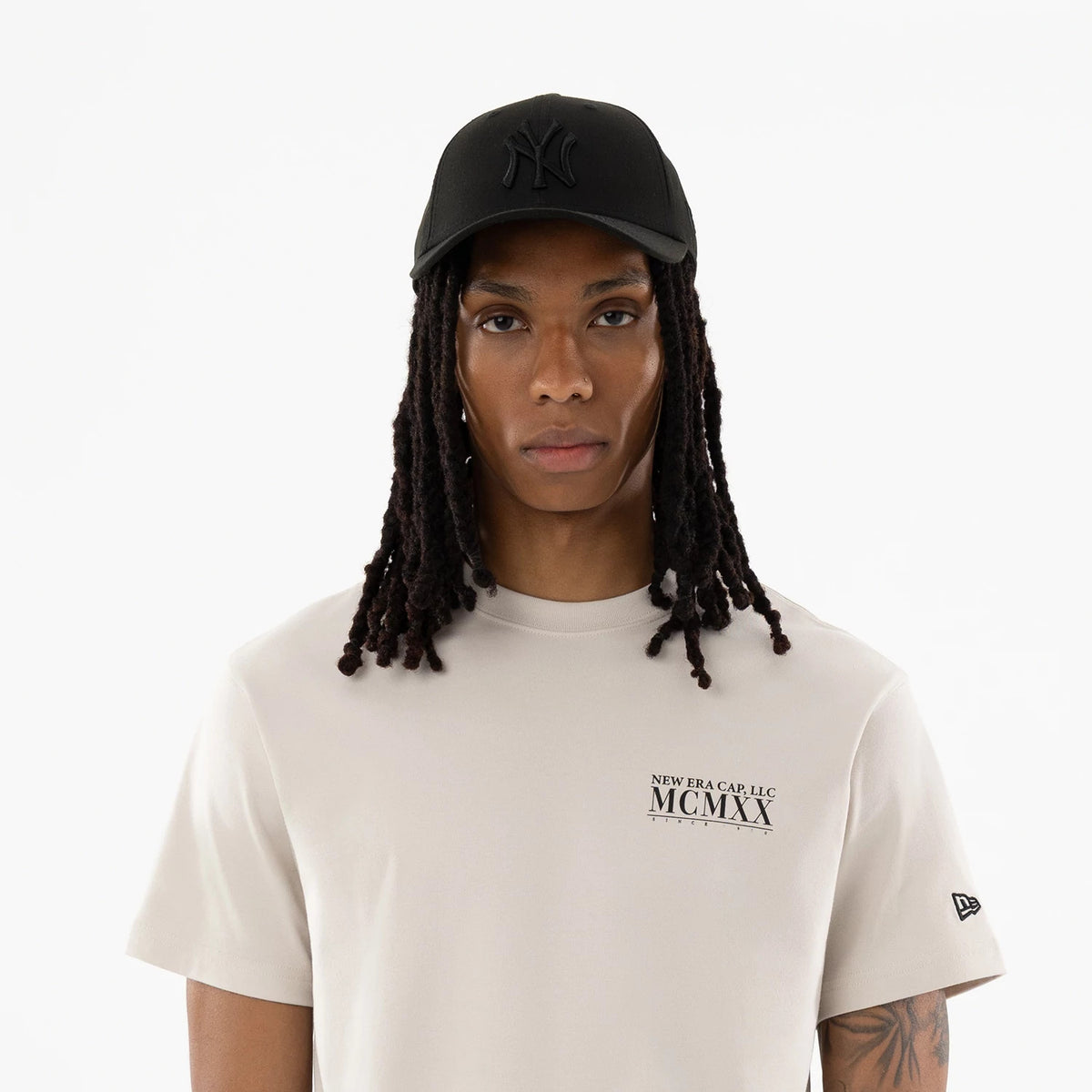 New Era Heritage Cream T-Shirt, on-model view showing the cream T-shirt with black text on the chest, a black tag logo on the left sleeve, paired with a black cap featuring a raised "NY" logo on the crown.