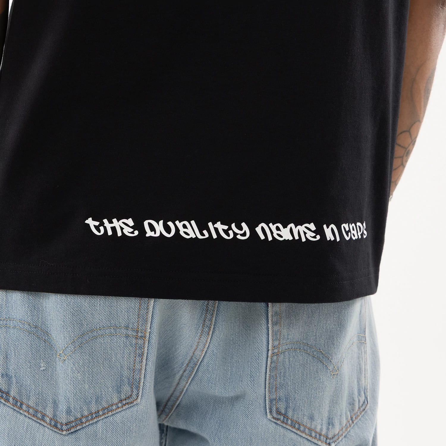 New Era I Am Original Black T-Shirt, on-model view showing the black fabric draping over light blue jeans, with off-white script text reading "the quality name in caps" near the bottom hem on the back.