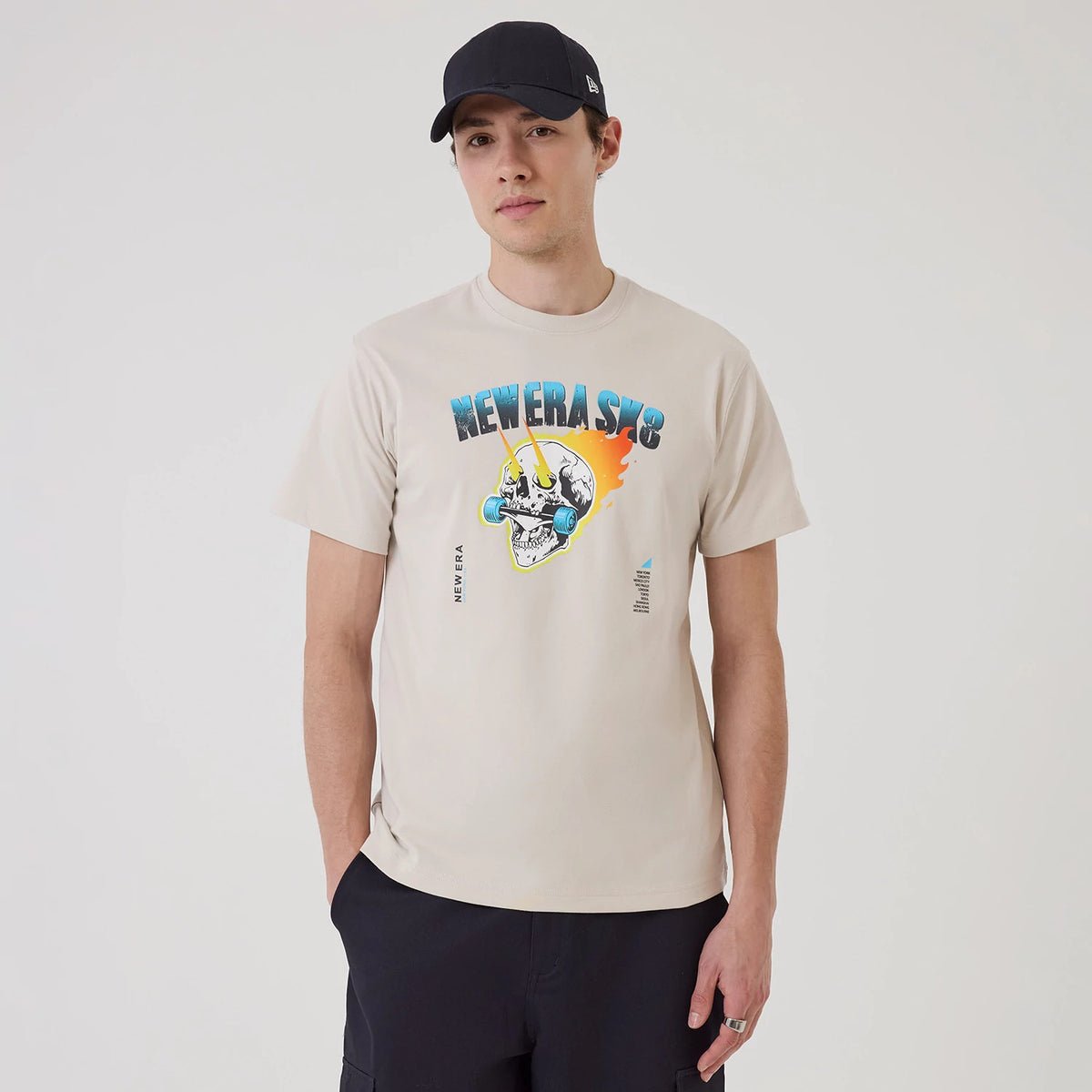 New Era SK8 Graphic Cream T-Shirt, front view showing a colourful graphic with 'NEWERA SK8' text above a skull with skateboards on the cream fabric, worn by a model in a black New Era cap.