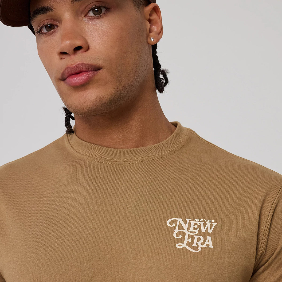 New Era Script Graphic Beige T-Shirt, on-model view showing a beige shirt with a raised white New Era script logo on the left chest, round neckline, and short sleeves worn by a person with a neutral expression.