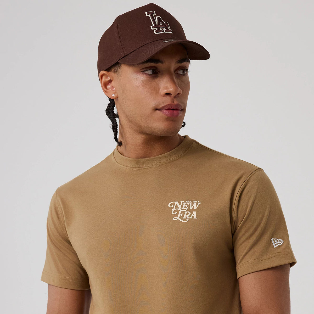 New Era Script Graphic Beige T-Shirt, front view showing the New Era logo printed in white on the left chest, a man wearing a brown cap with an embroidered LA logo, and a small New Era logo tag on the left sleeve.
