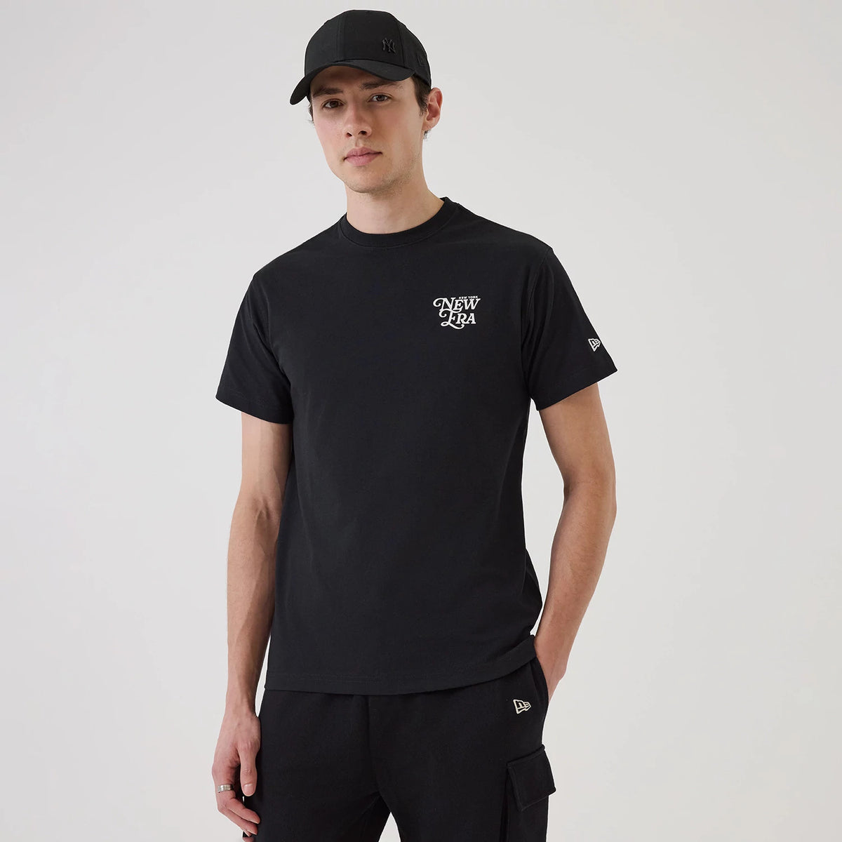 New Era Script Graphic Black T-Shirt, on-model view showing a black shirt with a white raised New Era logo on the left chest, short sleeves, and a debossed New Era logo on the left sleeve, paired with black trousers and a black cap.