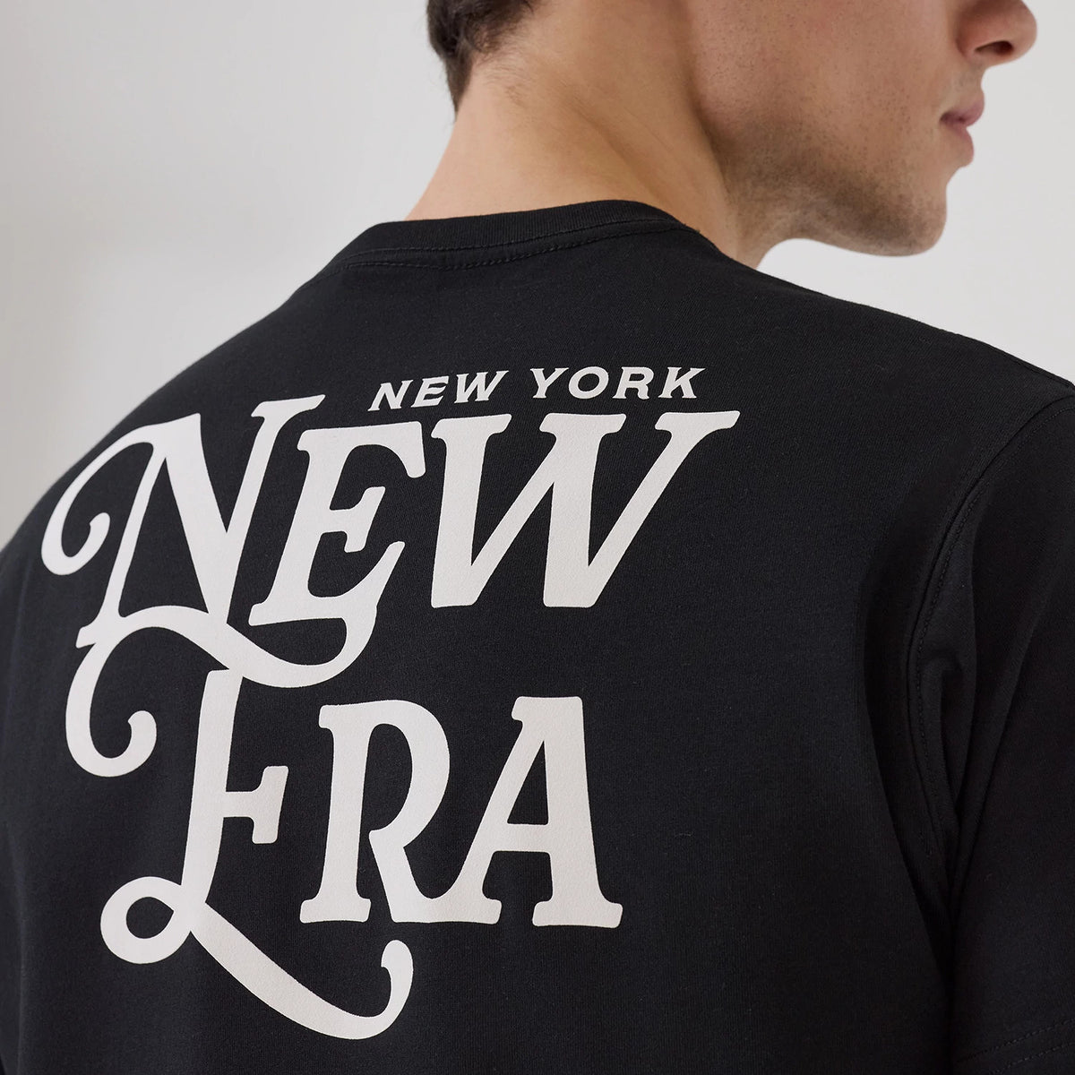 New Era Script Graphic Black T-Shirt, rear view showing a white script graphic featuring "New York New Era" centred on the upper back of the black fabric with part of the sleeve and neckline visible in profile.