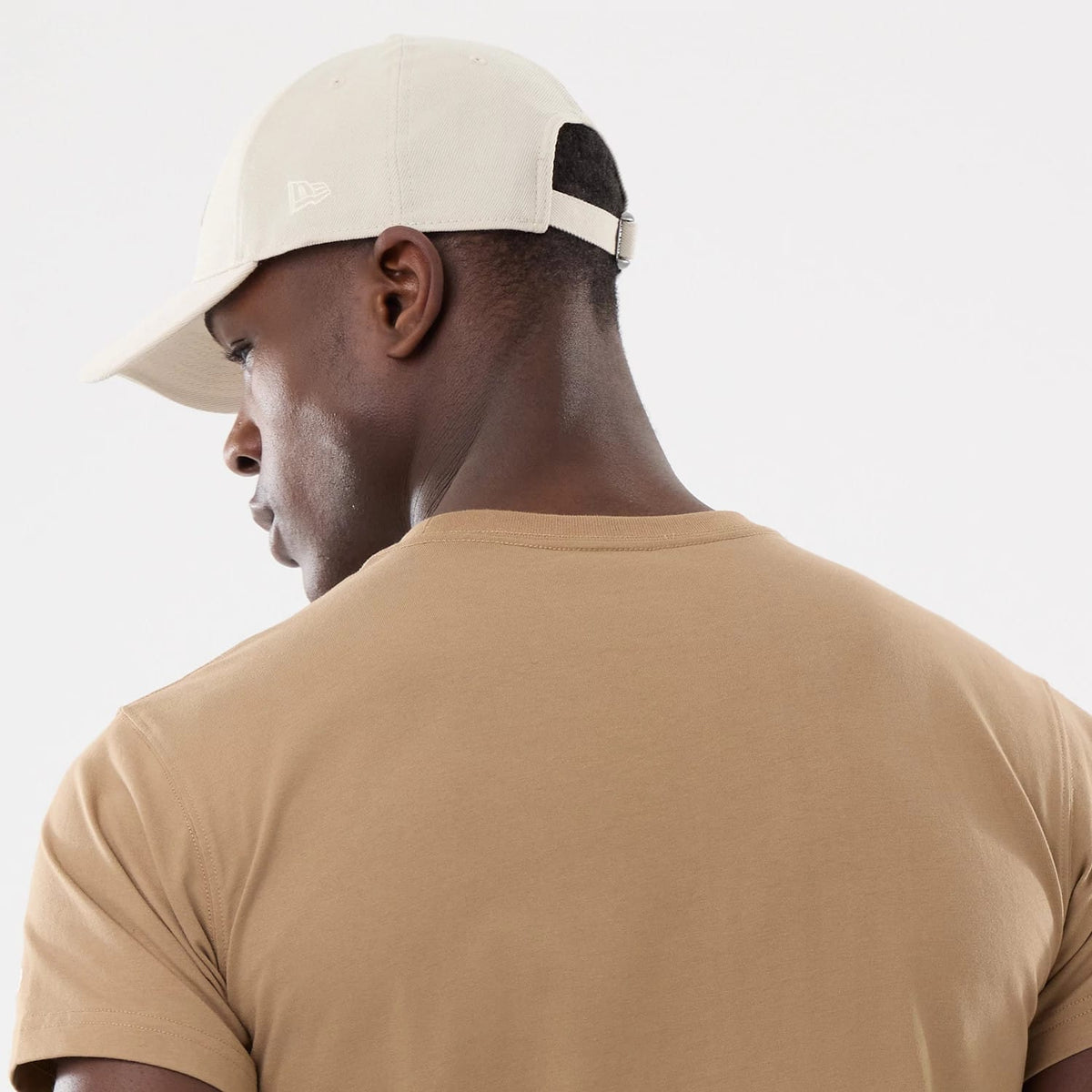 New Era Earth Day Beige T-Shirt, on-model view showing the back of the beige T-shirt against a beige cap with an embossed New Era logo on the left side, a black adjustable strap and eyelets near the top of the cap.