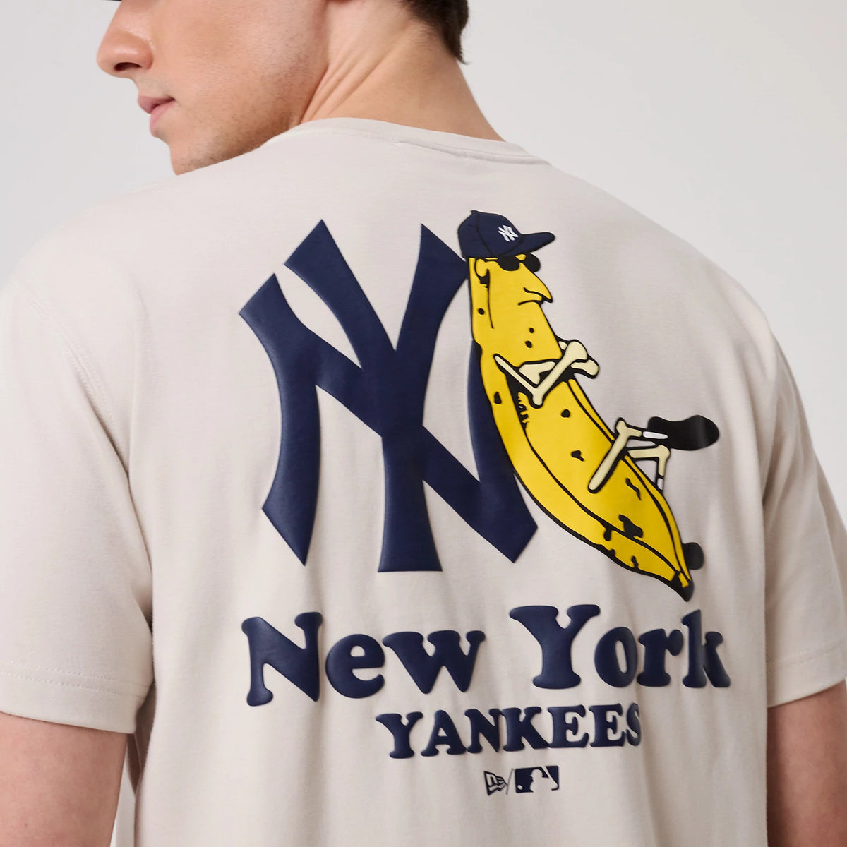 New York Yankees MLB Banana Graphic Natural T-Shirt, rear view showing a large navy New York Yankees logo with a yellow banana character graphic leaning on it, above bold blue lettering reading 'New York Yankees'.