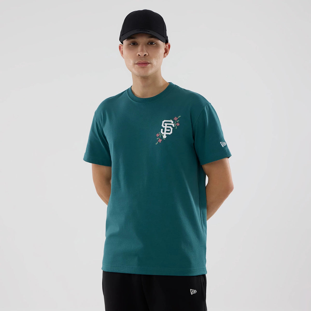 San Francisco Giants MLB Garden Dark Green T-Shirt, on-model view showing the dark green T-shirt with a raised SF logo and floral design on the left chest, New Era logo on the left sleeve, paired with black trousers and a black cap.