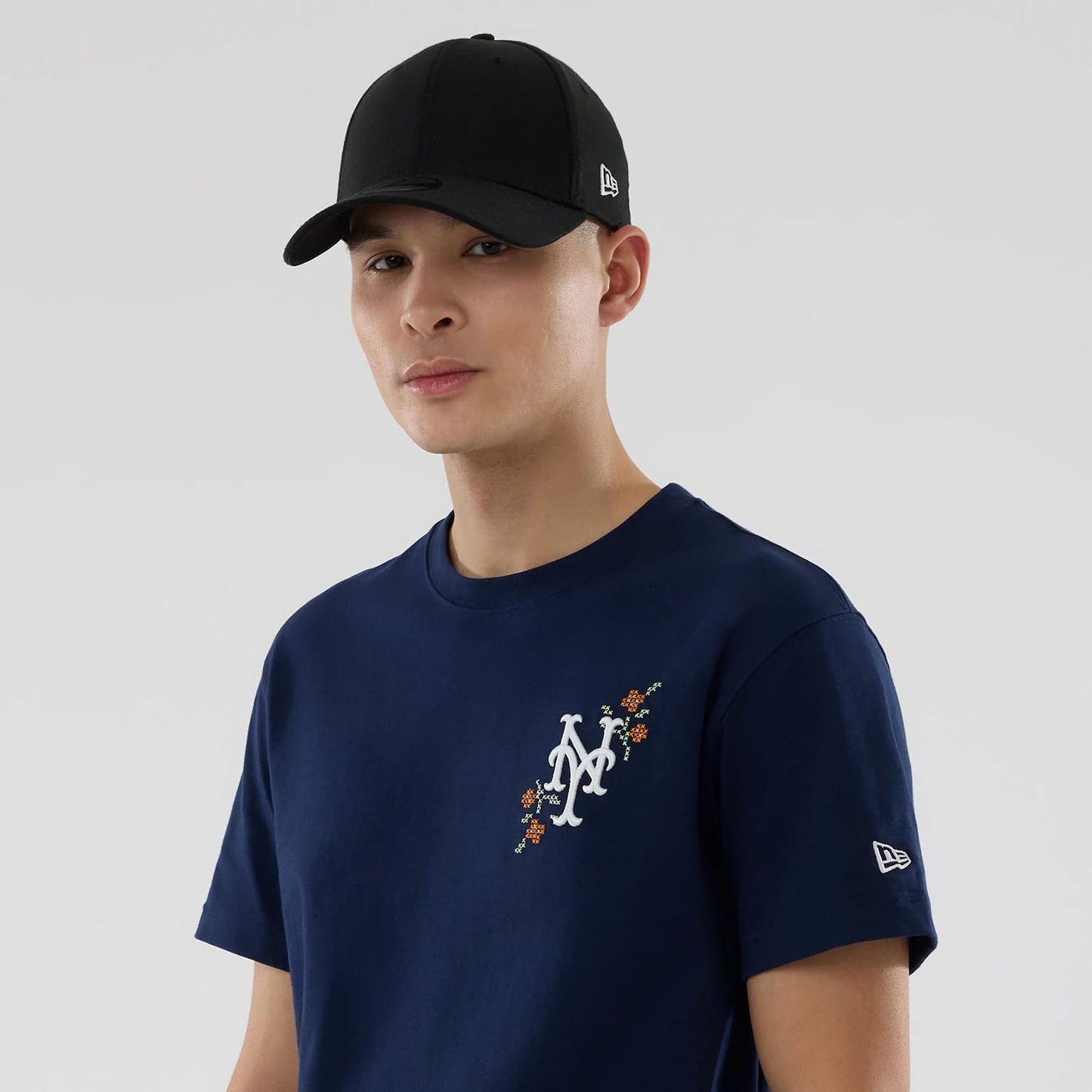 New York Mets MLB Garden Blue T-Shirt, on-model view showing the navy blue shirt with a white embroidered Mets logo framed by orange flowers on the chest and a raised New Era logo on the right sleeve, paired with a black cap.