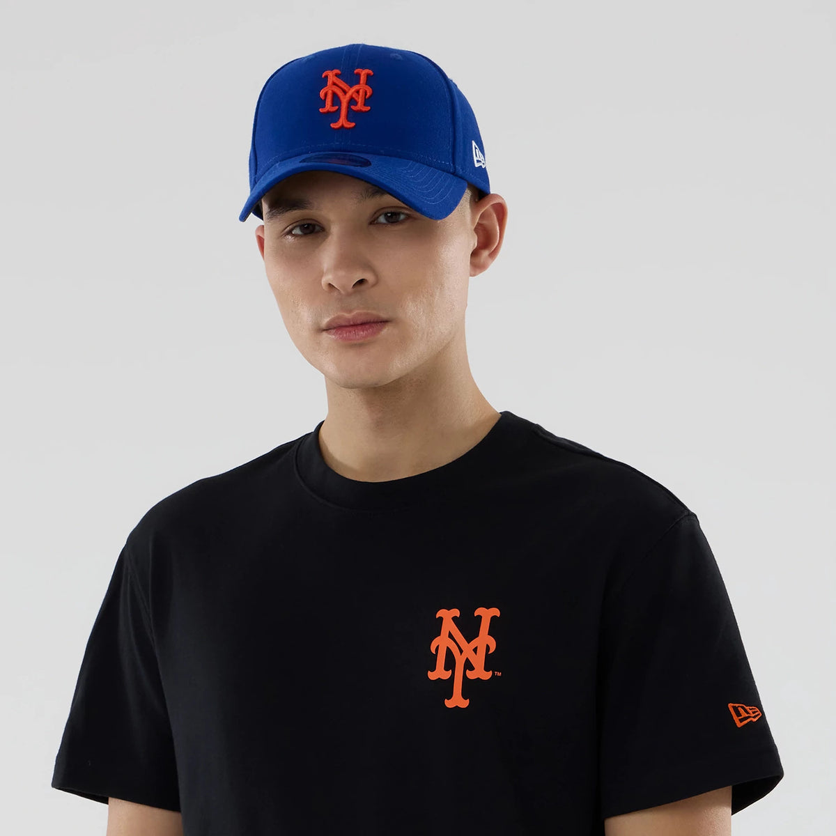 New York Mets MLB City Series Black T-Shirt, on-model view showing the orange New York Mets logo on the chest of the black T-shirt, a New Era logo on the sleeve, and a blue cap with an orange raised logo worn by the model.