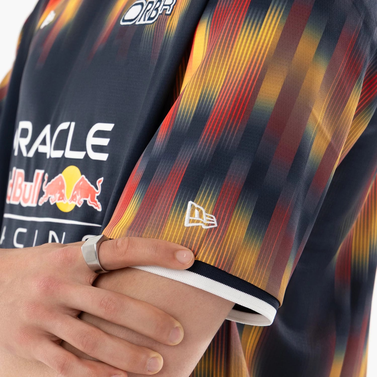 Red Bull Racing Multi Soccer Jersey, close-up on a model's folded arms showing the intricate gradient stripe pattern in red, yellow and navy on the sleeve, with the white embroidered New Era flag logo near the cuff.