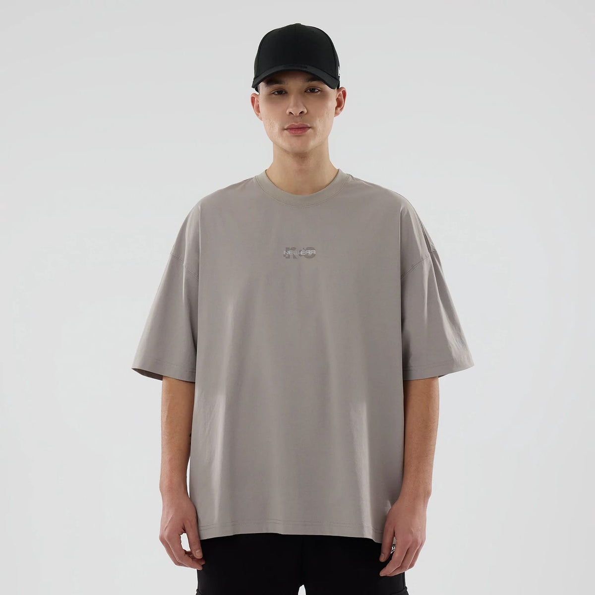 New Era NEC Logo Beige Oversized T-Shirt, front view showing a debossed NEC logo centred on the chest, loose short sleeves and a relaxed fit design worn by a person in black trousers.