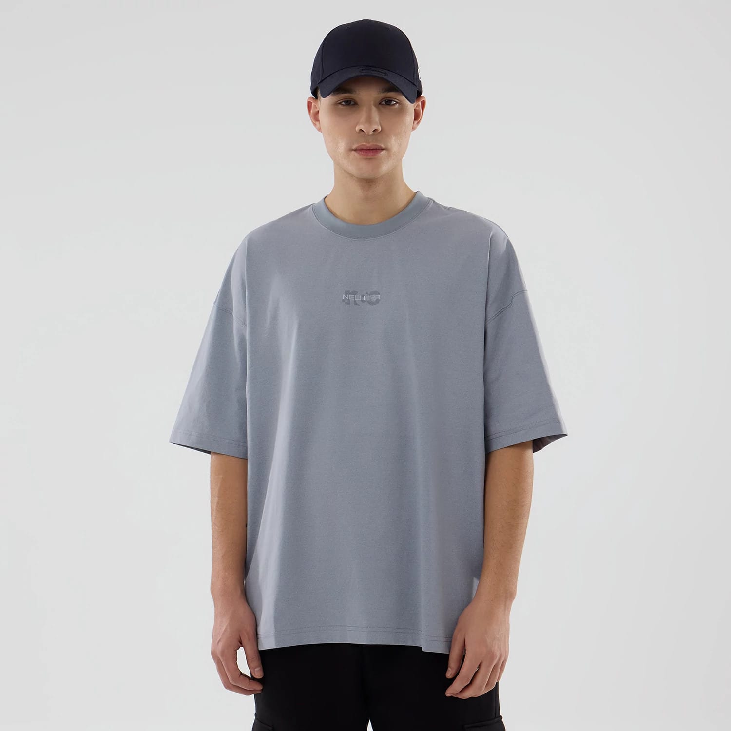 New Era NEC Logo Grey Oversized T-Shirt, front view showing a light grey oversized fit with a debossed NEC logo centred on the chest, worn by a model pairing it with a plain dark cap and black trousers.