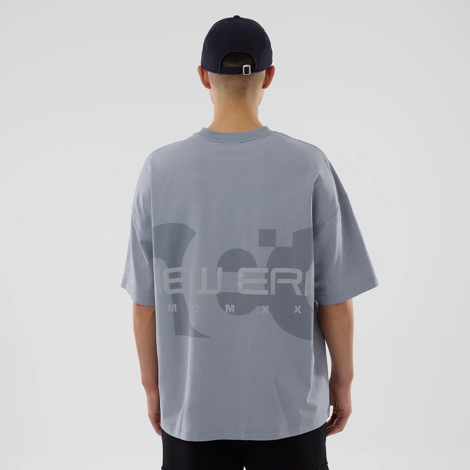 New Era NEC Logo Grey Oversized T-Shirt, on-model rear view showing the oversized grey fit with a large printed "NEW ERA MMXX" logo across the upper back, worn by a person in a black cap.