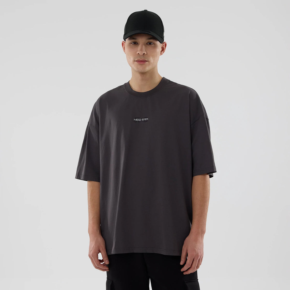 New Era NEC Logo Black Oversized T-Shirt, front view showing a charcoal grey oversized T-shirt with a small white debossed New Era logo centred on the chest, worn by a person standing in neutral lighting.