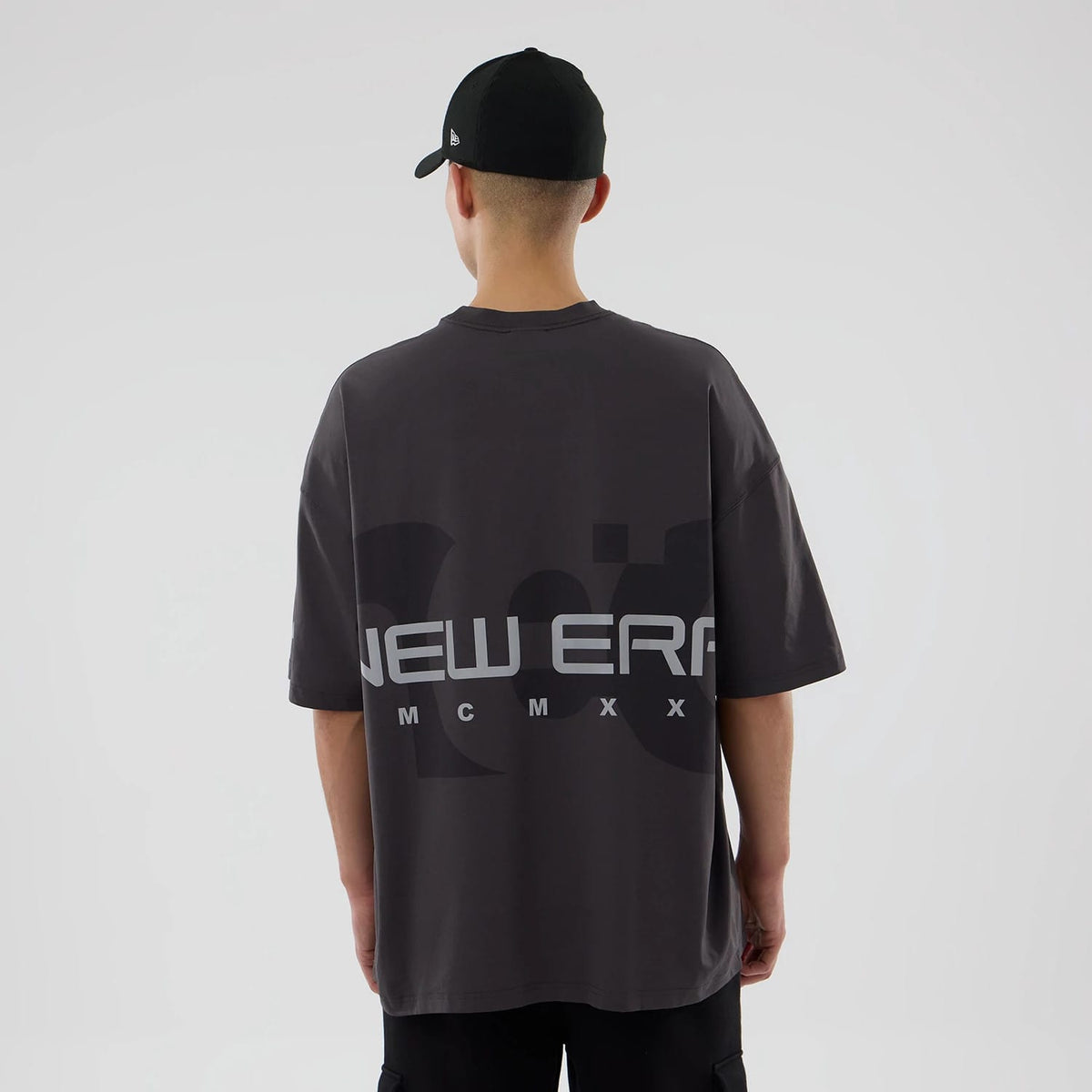 New Era NEC Logo Black Oversized T-Shirt, rear view showing a large logo design featuring the words "NEW ERA" and "MCMXX" in grey across the back, worn by a person in a black cap with a debossed logo on the side.