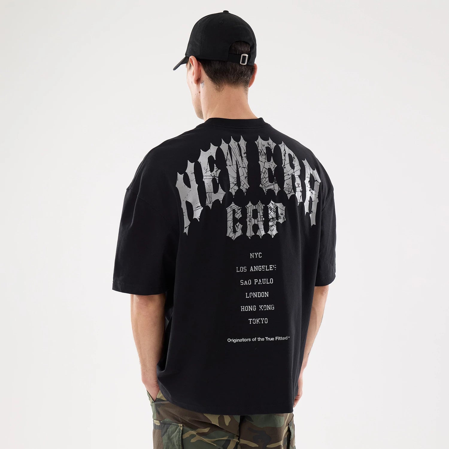 New Era Graphic Street Archived City Black Oversized T-Shirt, rear view with large gothic grey text reading "New Era Cap" above city names on black fabric, worn by a person in camo trousers and a black cap.