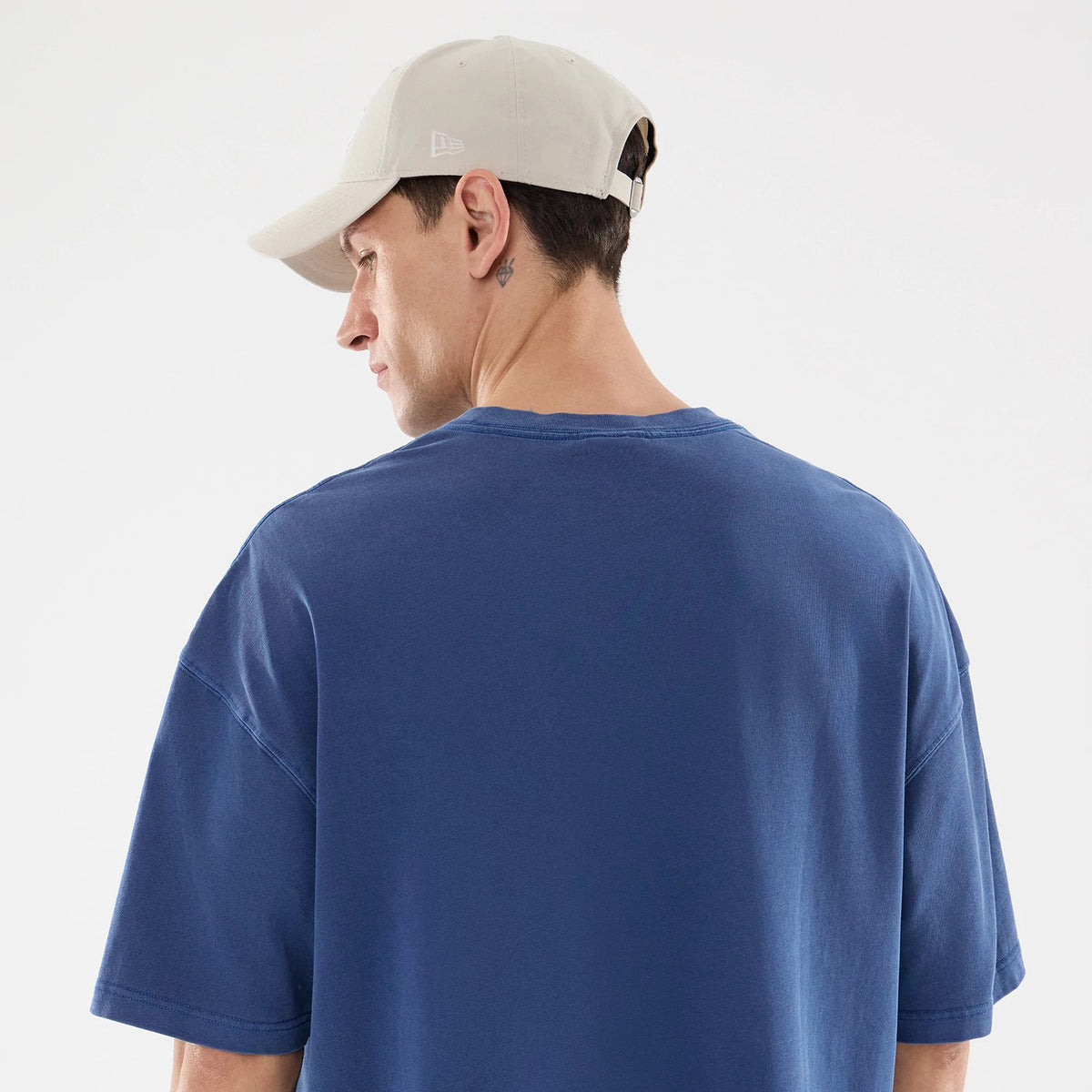 New Era Graphic Street Archived Blue Oversized T-Shirt, on-model rear view highlighting the plain blue back with short sleeves, worn by a person in a beige cap with a raised New Era logo on the side.
