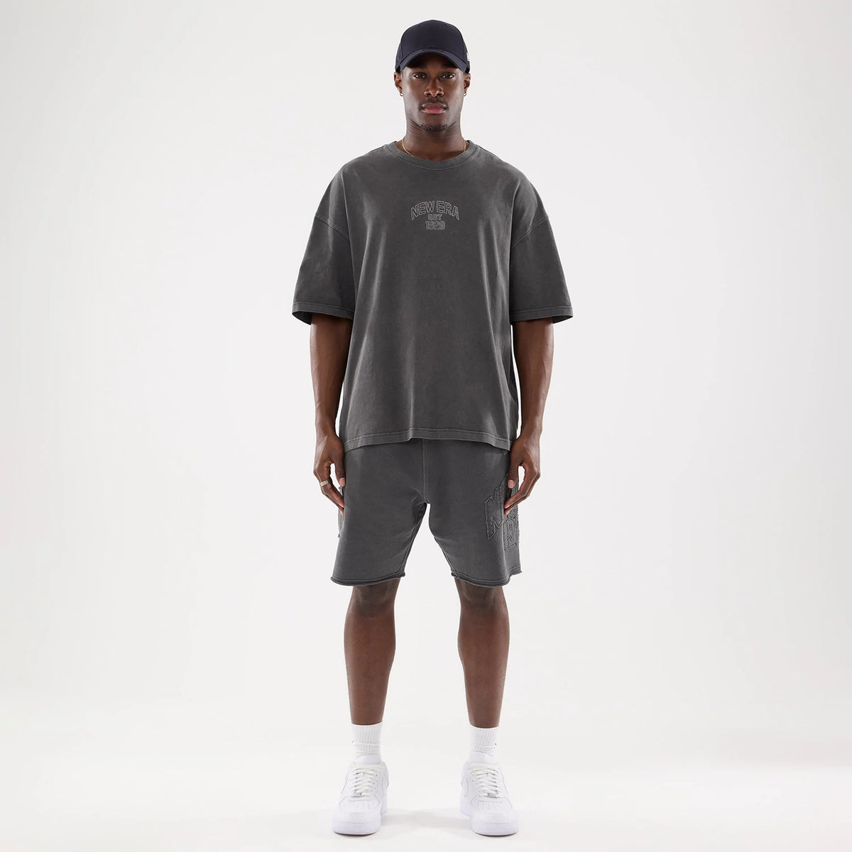 New Era Graphic Street Archived Washed Black Oversized T-Shirt, front view showing an embossed New Era logo centred on the chest of the faded black oversized tee, worn with matching shorts by a model in white sneakers and a black cap.