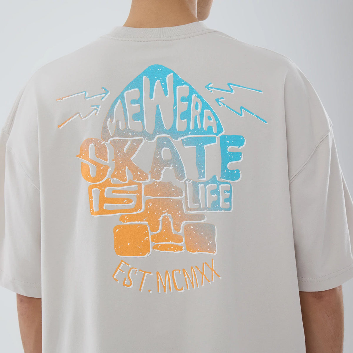New Era Skate Is Life Graphic Cream Oversized T-Shirt, back view showing a large graphic in blue and orange with the text "New Era Skate Is Life EST. MCMXX" in a stylized font across the back on a cream fabric.