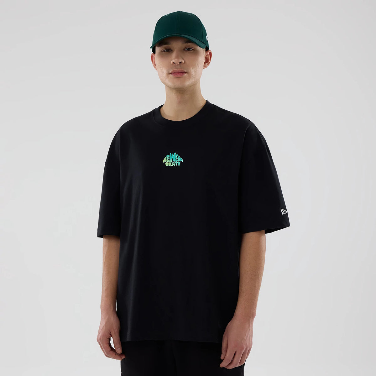 New Era Skate Is Life Graphic Black Oversized T-Shirt, on-model view displaying a black tee with a green "SKATE" graphic on the chest and a debossed white New Era logo on the left sleeve, worn with a green cap.