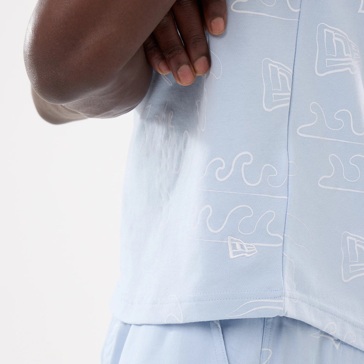 New Era Resort Blue Tank Top, close-up showing the side seam, featuring an all-over pattern of white wave graphics and New Era flag logos, with soft blue fabric and stitching details near the hem and armhole.