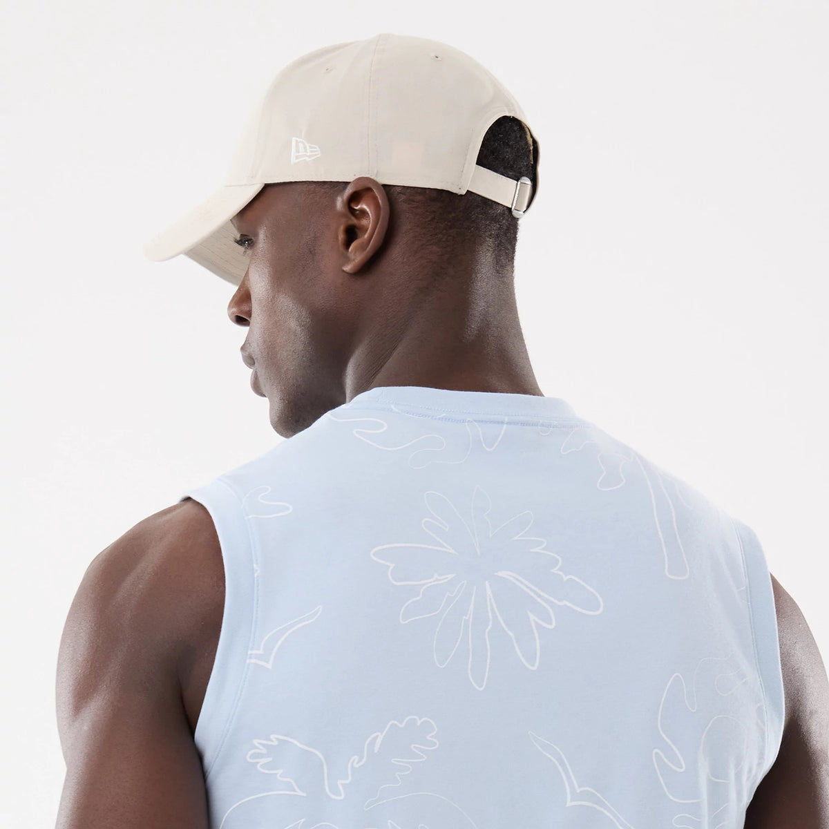 New Era Resort Blue Tank Top, on-model view showing white outline palm illustrations on the back, paired with a beige cap featuring the New Era flag logo on the left side and a metal buckle strap.