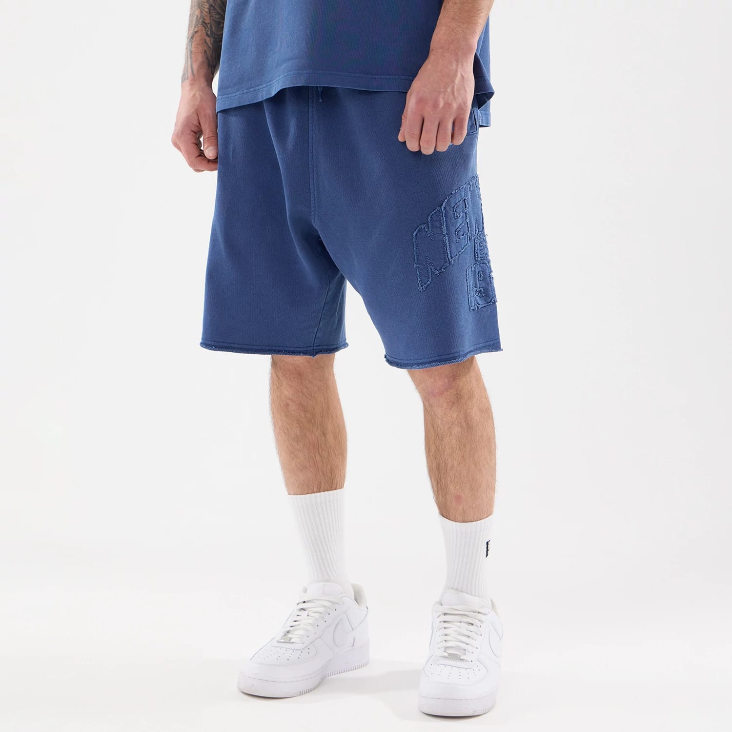 New Era Street Archived Blue Oversized Shorts, on-model view showing the knee-length blue shorts with an embossed New Era logo on the left leg, featuring a drawstring waistband and a loose fit.